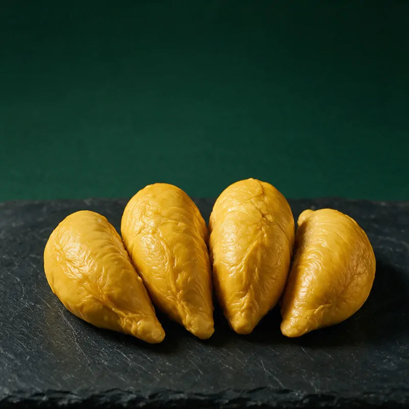 Four whole lobes of golden Musang King durian pulp arranged on a dark slate slab against a deep green studio wall.